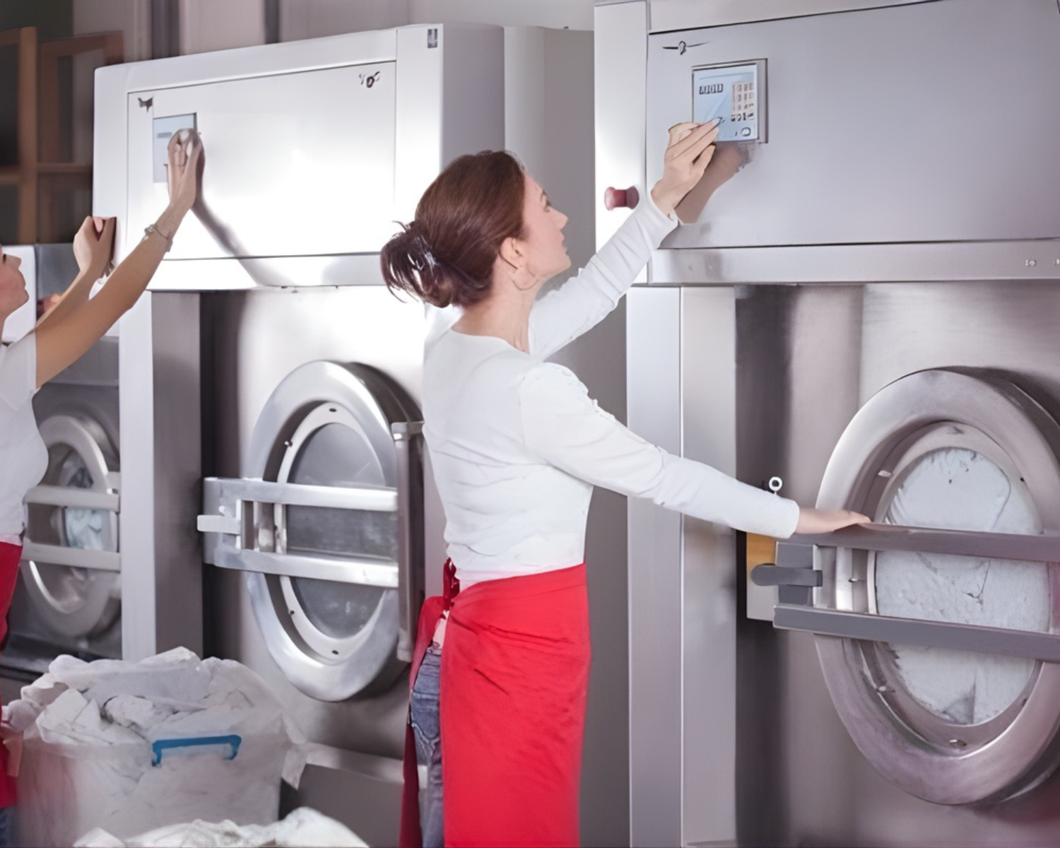 Staff loading and operating large industrial washing machines in a commercial laundry room.
