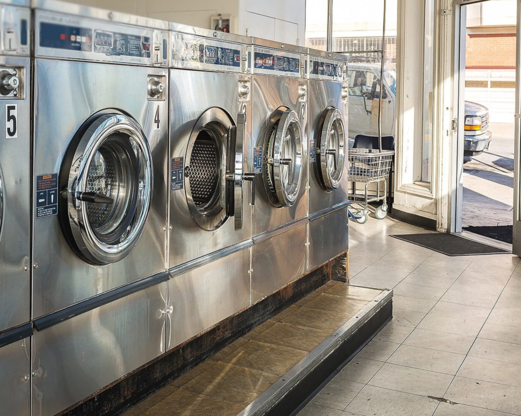 Row of stainless steel commercial washers inside a bright laundromat, representing equipment solutions supported by Southeastern Laundry