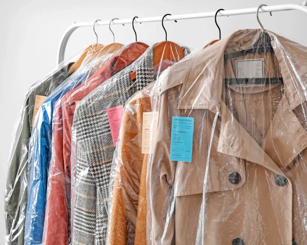 Garments in protective covers hanging on a rack at a dry cleaner facility supported by commercial laundry equipment in Greenwood, SC.